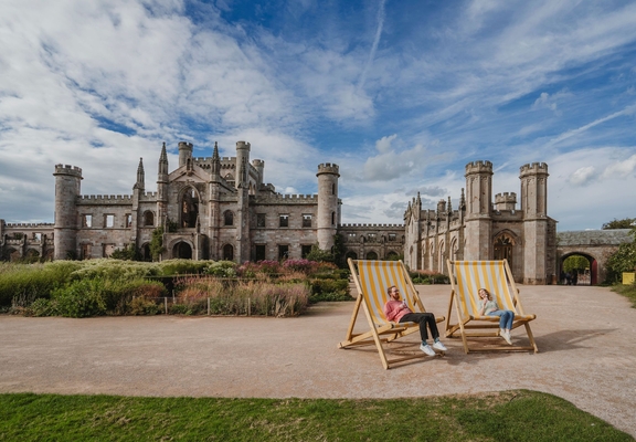 Man and woman holding ice creams in oversized deck chairs in manicured castle grounds and gardens.
