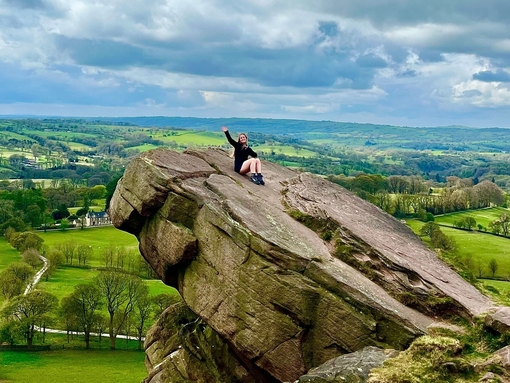 A seated woman waving from a hanging stone, overlooking a country panorama.