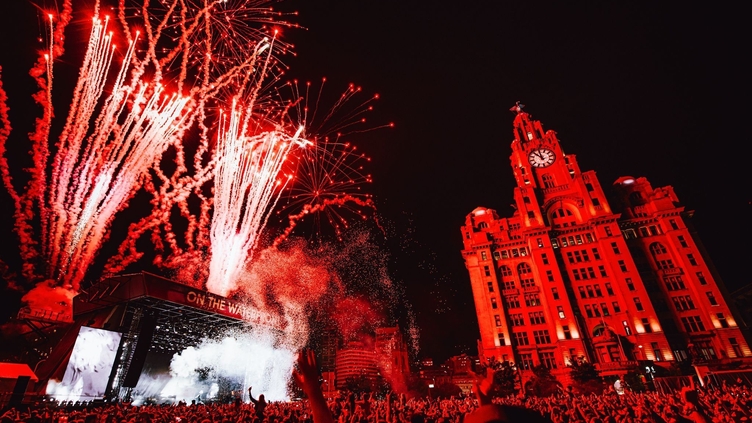 Crowds of people watching a band at a music festival as fireworks explode, in front of the Liver Building.