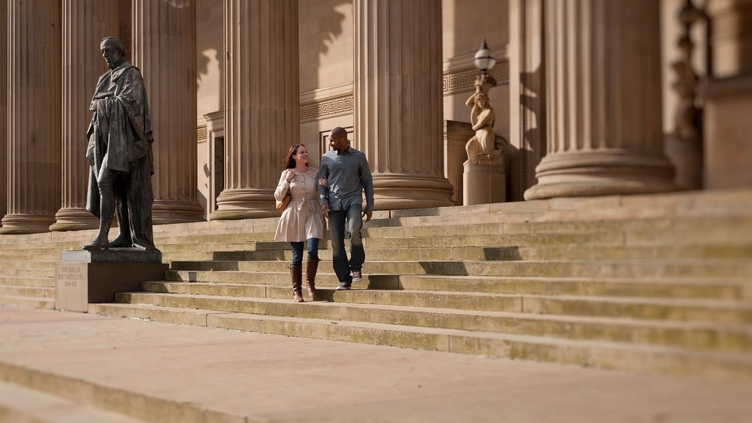 St George's Hall on Lime Street in Liverpool city centre. A neoclassical building, with a statue of Disraeli on the steps. A couple walking past the building.