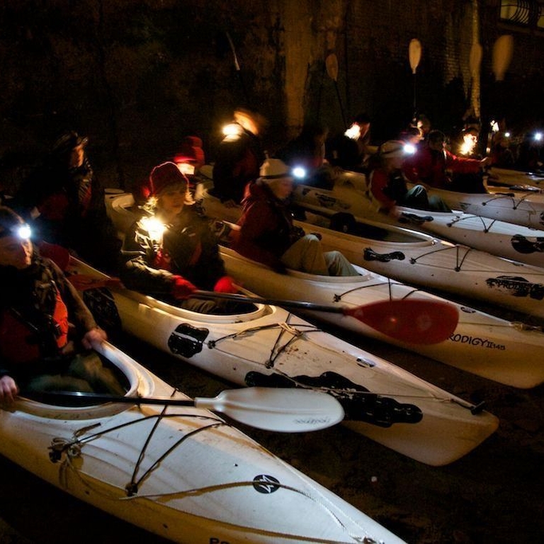 Row of people with head torches sat waiting in white canoes in water ready to go out for the night tour