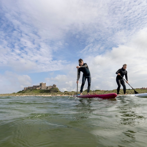 Two men on are paddleboarding in the sea with a heritage castle in the background.