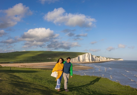 A couple walk together on a headland with white chalk cliffs in the background.