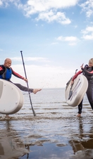 A man and woman in wet suits stand holding paddleboards on a beach
