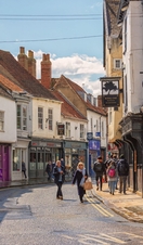 Shopping street just after a rain shower. Historic buildings line the street and shoppers walk between them. A sky with cloud is overhead.