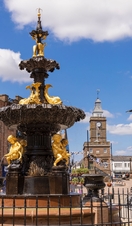An ornate black and gold fountain with statues of people and fish in the centre of a shopping street.