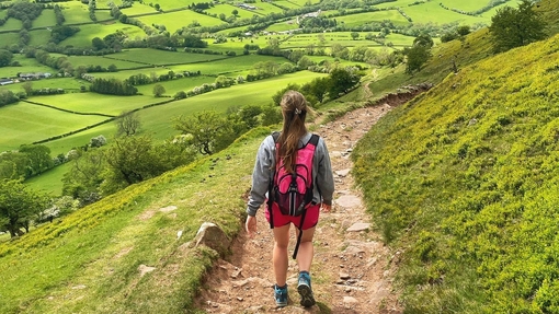 Rückenansicht einer jungen Frau, die auf einem Weg durch ein üppig grünes Tal und über Hügel geht
