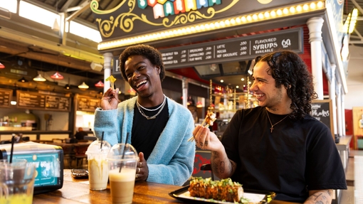 Two friends laugh while eating and drinking inside a food market