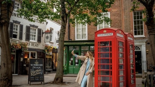 A woman leans against a red telephone box and takes a selfie in a town square.