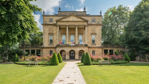Summertime view of a heritage house set in gardens with topiary bushes and paved path entrance.