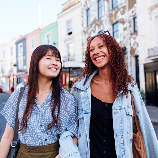 Two young women linking arms and walking in a street smiling