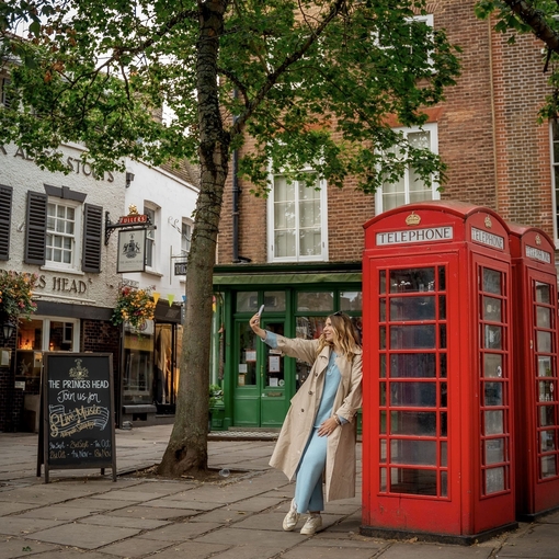 A woman leans against a red telephone box and takes a selfie in a town square.