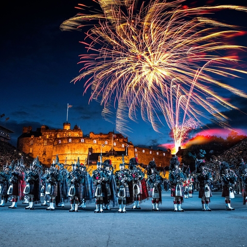 The arena at the Edinburgh Military Tattoo during a performance of the military event, parade ground and packed spectator seating. A light show projecting onto the castle walls. Marching band with a leading conductor, and massed pipers playing the bagpipes. Fireworks exploding in the night sky.