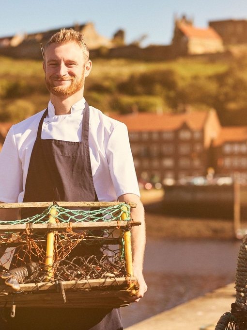Chef wearing apron on pier holding lobster trap with lobster