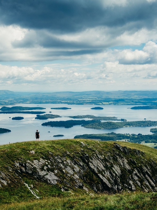 A wide panorama of a hiker standing atop a hill looking out over grassland and lakes.
