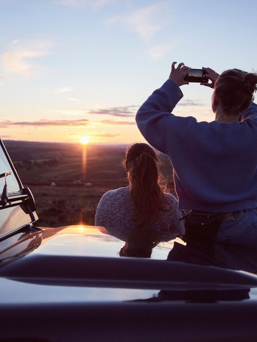 Women sitting on bonnet of 4x4 parked on road at sunset