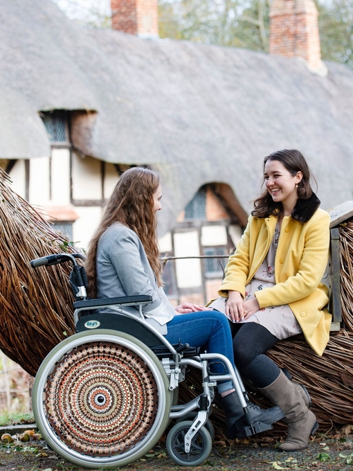 Rollstuhlfahrer und Freund sitzen neben einer Holzskulptur