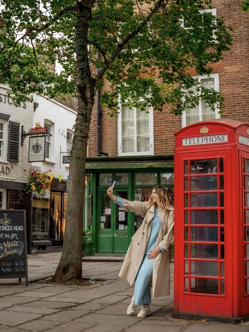 A woman leans against a red telephone box and takes a selfie in a town square.