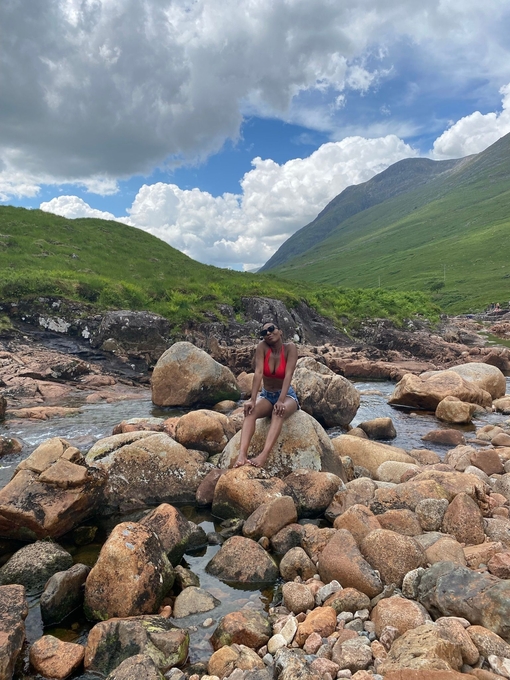 A woman sits on a rock surrounded by a stream and hills beyond