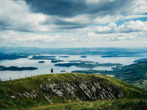 A wide panorama of a hiker standing atop a hill looking out over grassland and lakes.
