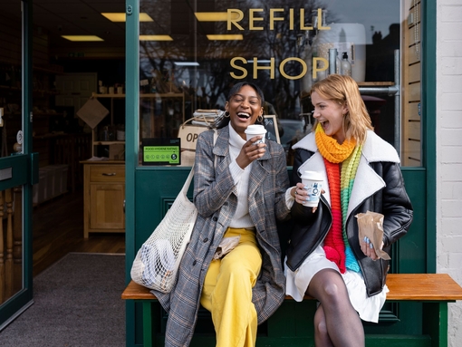 Two young women sat on a bench outside a coffee shop with takeaway coffees