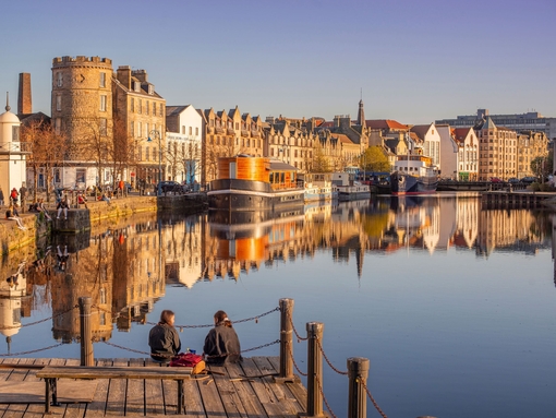 Two young women sat on the jetty of the waterfront in the evening with boats and buildings by the harbour.