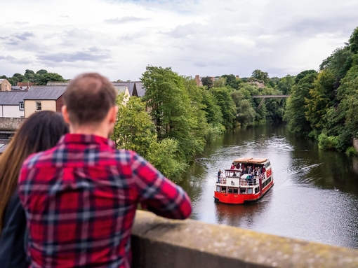 Two people stood on a bridge overlooking a tour boat sailing down a river in Durham