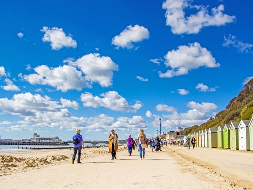 People walking along the coast next to colourful beach huts on a sunny day.