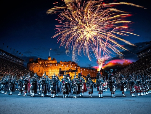 The arena at the Edinburgh Military Tattoo during a performance of the military event, parade ground and packed spectator seating. A light show projecting onto the castle walls. Marching band with a leading conductor, and massed pipers playing the bagpipes. Fireworks exploding in the night sky.