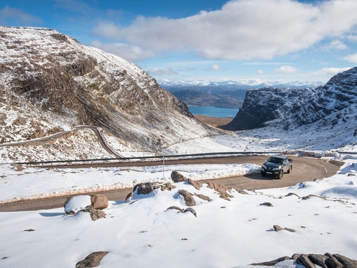 A pickup truck drives up a windy, hilly road lined with snow in winter
