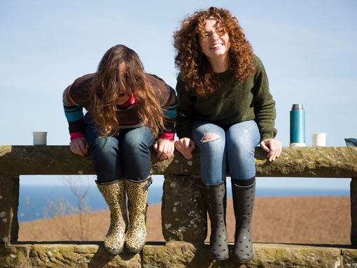 Two cheerful women holding mugs with hot drinks, laughing