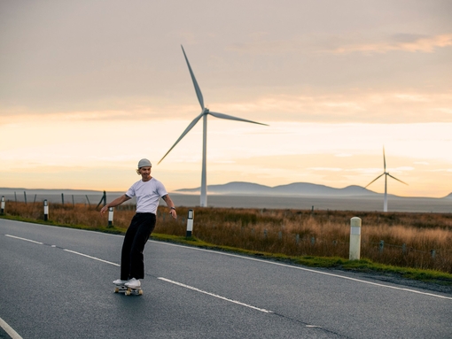 A male skateboarder enjoys the flat road surface to skate on in the Scottish Highlands, with wind turbines behind him
