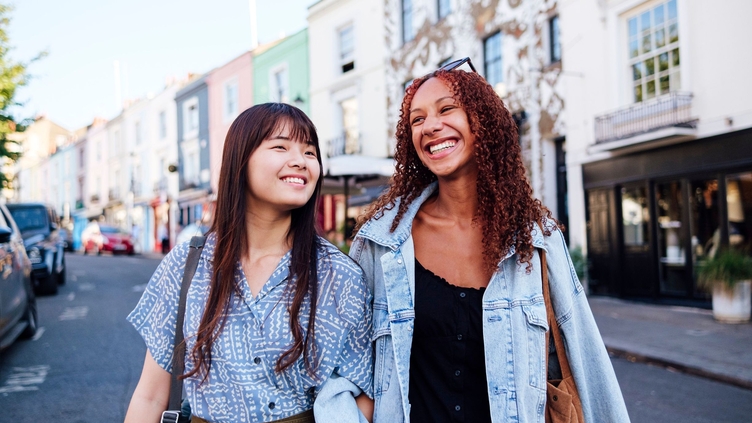 Two young women linking arms and walking in a street smiling