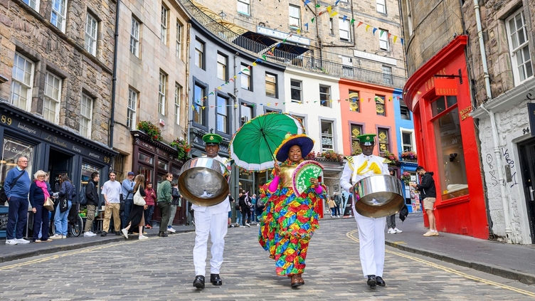 Festival performers in traditional dress walking down a street carrying steel drums and a parasol