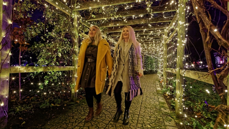 Two women walking through a wooden arched walkway lit up with fairy lights.