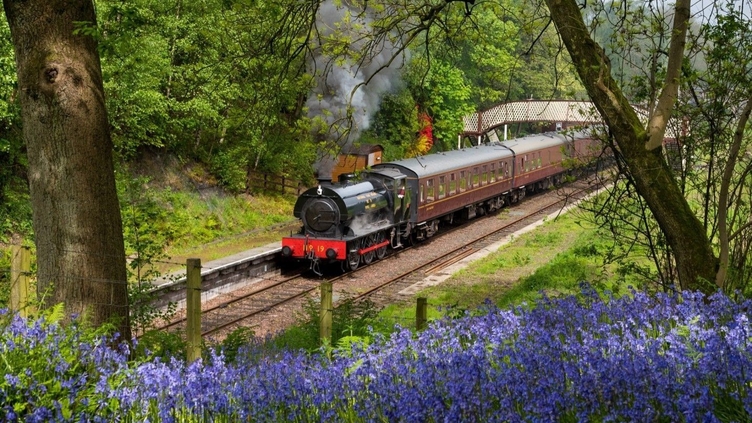 A steam train with lots of steam coming out, pulling at least three vintage carriages, gets ready to depart a small traditional railway station.