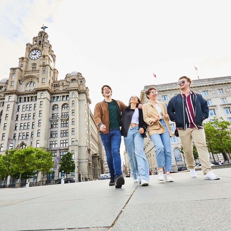 أربعة أصدقاء يستكشفون مدينة محلية وهم يتمشون على ممشى الواجهة البحرية، وخلفهم مبان تاريخية.